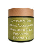 A jar of essential oil blend with a light wood-grain lid and a green label reading 'Grass-fed Beef Tallow, Avocado Oil, Therapeutic Grade Peppermint Essential Oil' on a white background.