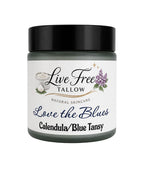 A jar of Live Free Tallow Natural Skincare 'Love the Blues' Calendula/Blue Tansy cream, featuring a white label with black and blue text and illustrations, and a black lid, on a white background.